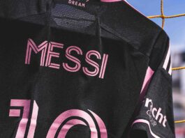 A black jersey with pink writing reads “Messi 10” on the back. In the background, a soccer field can be seen.