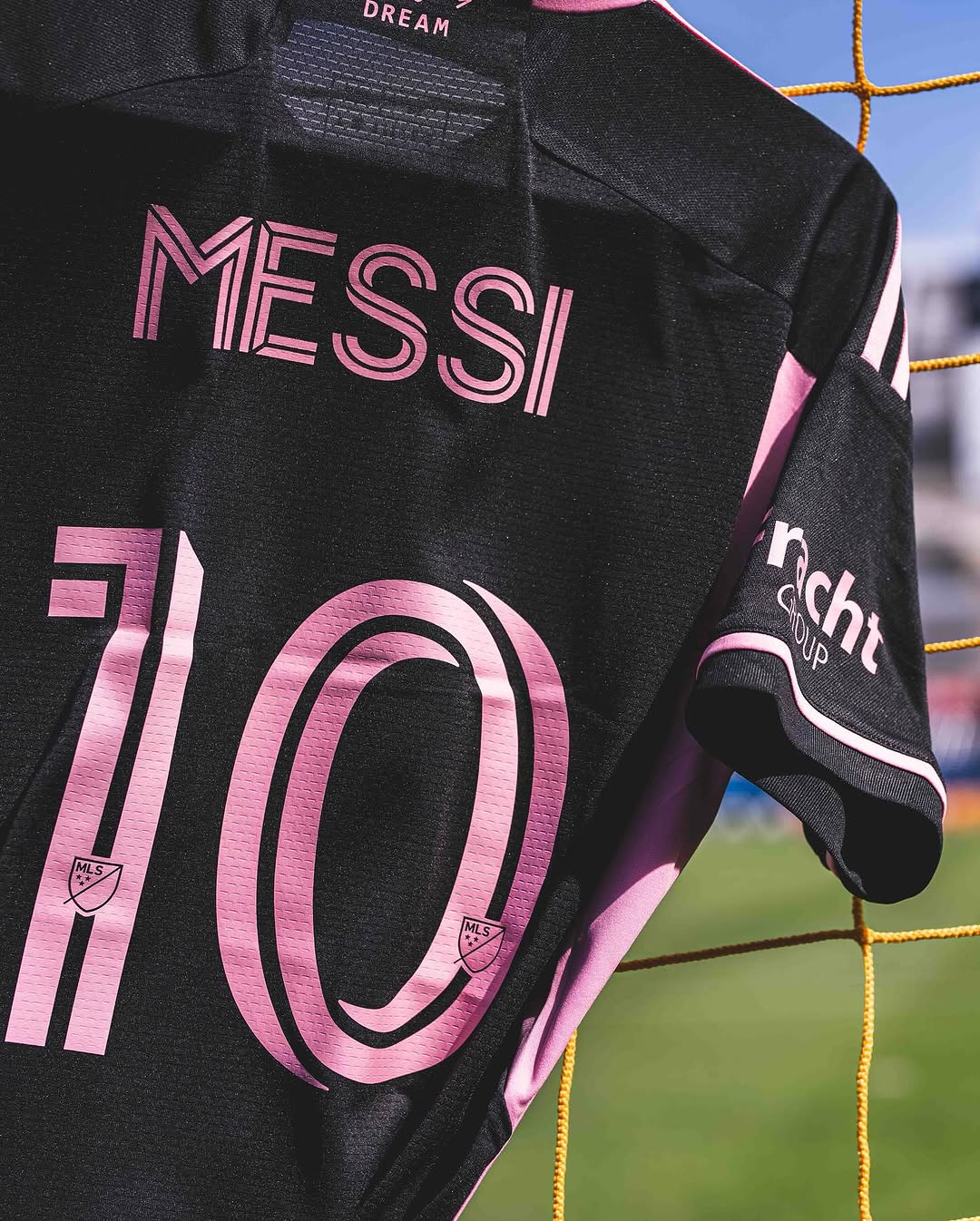 A black jersey with pink writing reads “Messi 10” on the back. In the background, a soccer field can be seen.