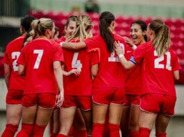 Members of the women’s soccer team, wearing red uniforms with white lettering, celebrate with a group embrace.