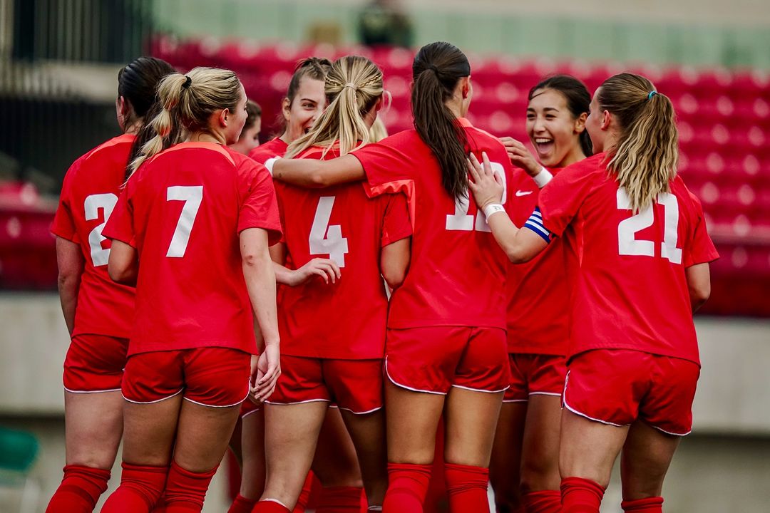 Members of the women’s soccer team, wearing red uniforms with white lettering, celebrate with a group embrace.