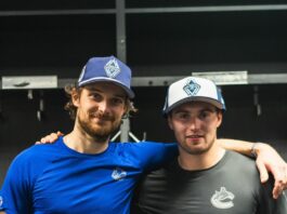 Two men wearing shirts with the Canucks orca logo pose with baseball caps decorated with the Whitecaps logo resembling mountains.