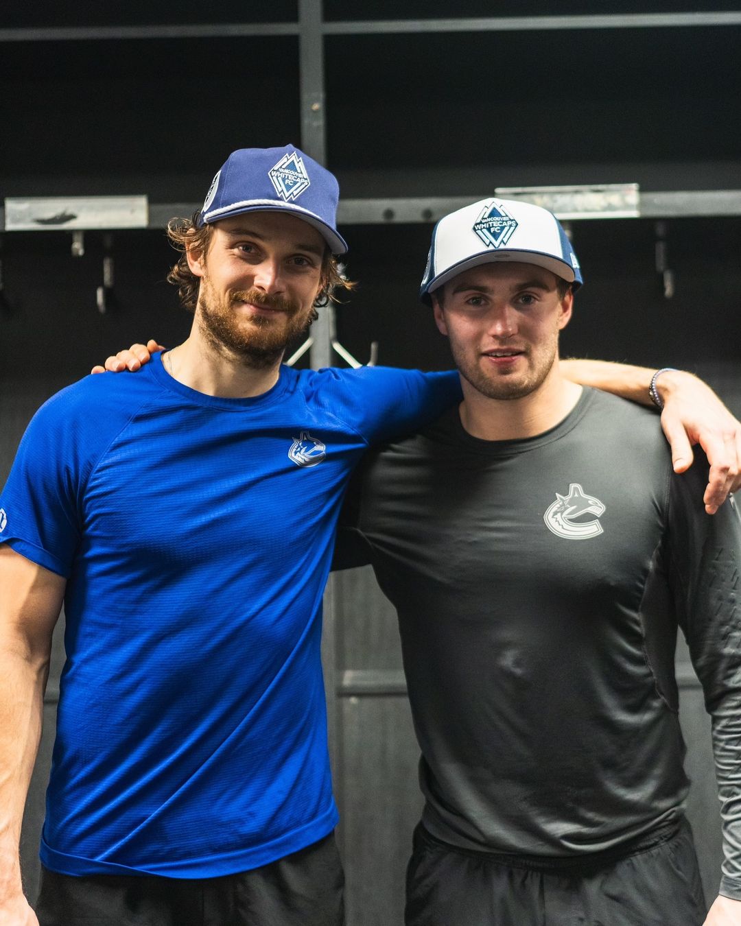 Two men wearing shirts with the Canucks orca logo pose with baseball caps decorated with the Whitecaps logo resembling mountains.