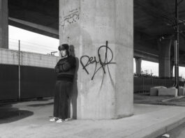 In black and white, under a concrete bridge, a boy stands in a sweater and jeans as he leans against a pillar.