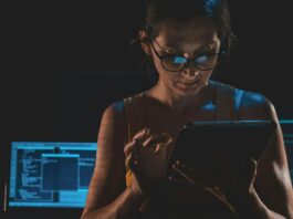 Woman standing in a dark room scrolling on a tablet. Behind her, there are multiple computer screens with code on them.