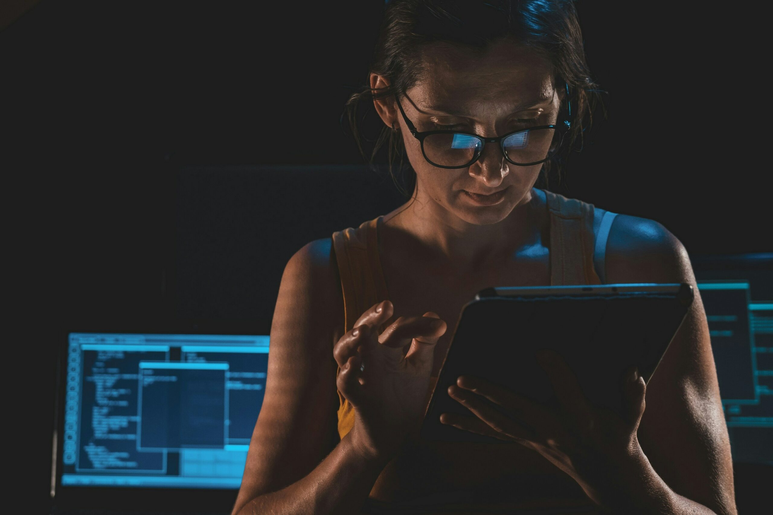 Woman standing in a dark room scrolling on a tablet. Behind her, there are multiple computer screens with code on them.