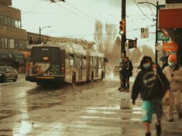 99 B-Line bus crossing an intersection on a rainy day. There are pedestrians wearing masks who are crossing as well.