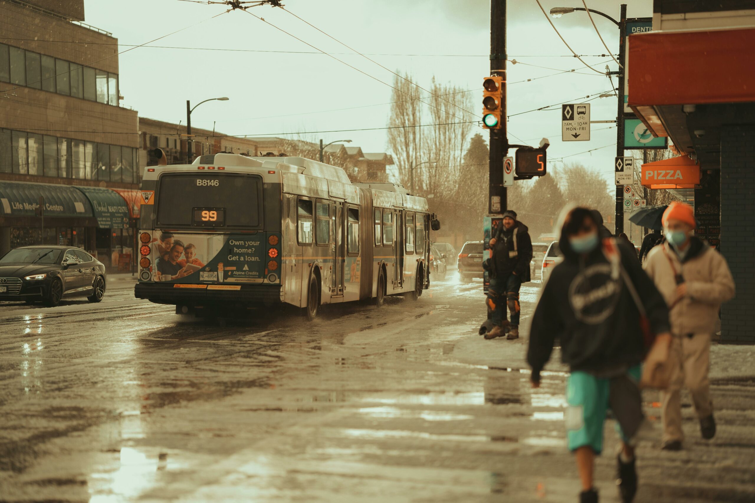 99 B-Line bus crossing an intersection on a rainy day. There are pedestrians wearing masks who are crossing as well.