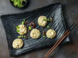 Five circular dumplings with pinched tops sit on a black plate garnished with green sprouts.