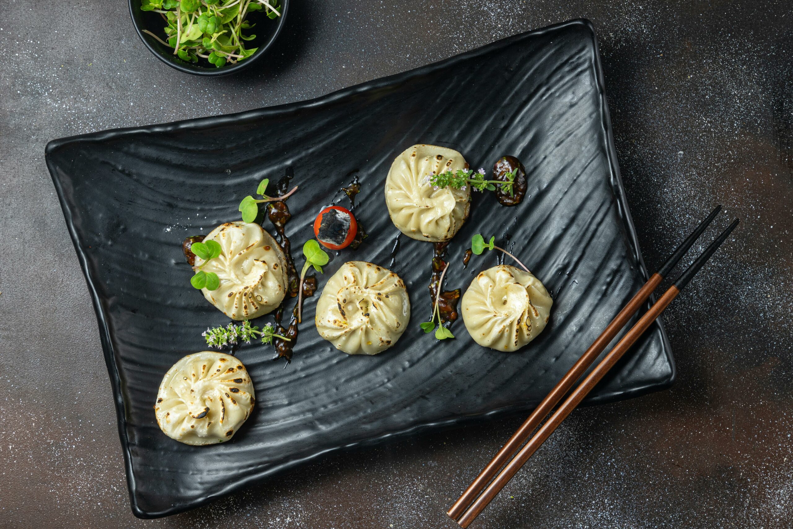 Five circular dumplings with pinched tops sit on a black plate garnished with green sprouts.