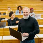 It’s Dear Peakie again Professor standing in front of a lecture hall. He is holding a laptop and smiling at the camera.