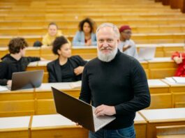 Professor standing in front of a lecture hall. He is holding a laptop and smiling at the camera.