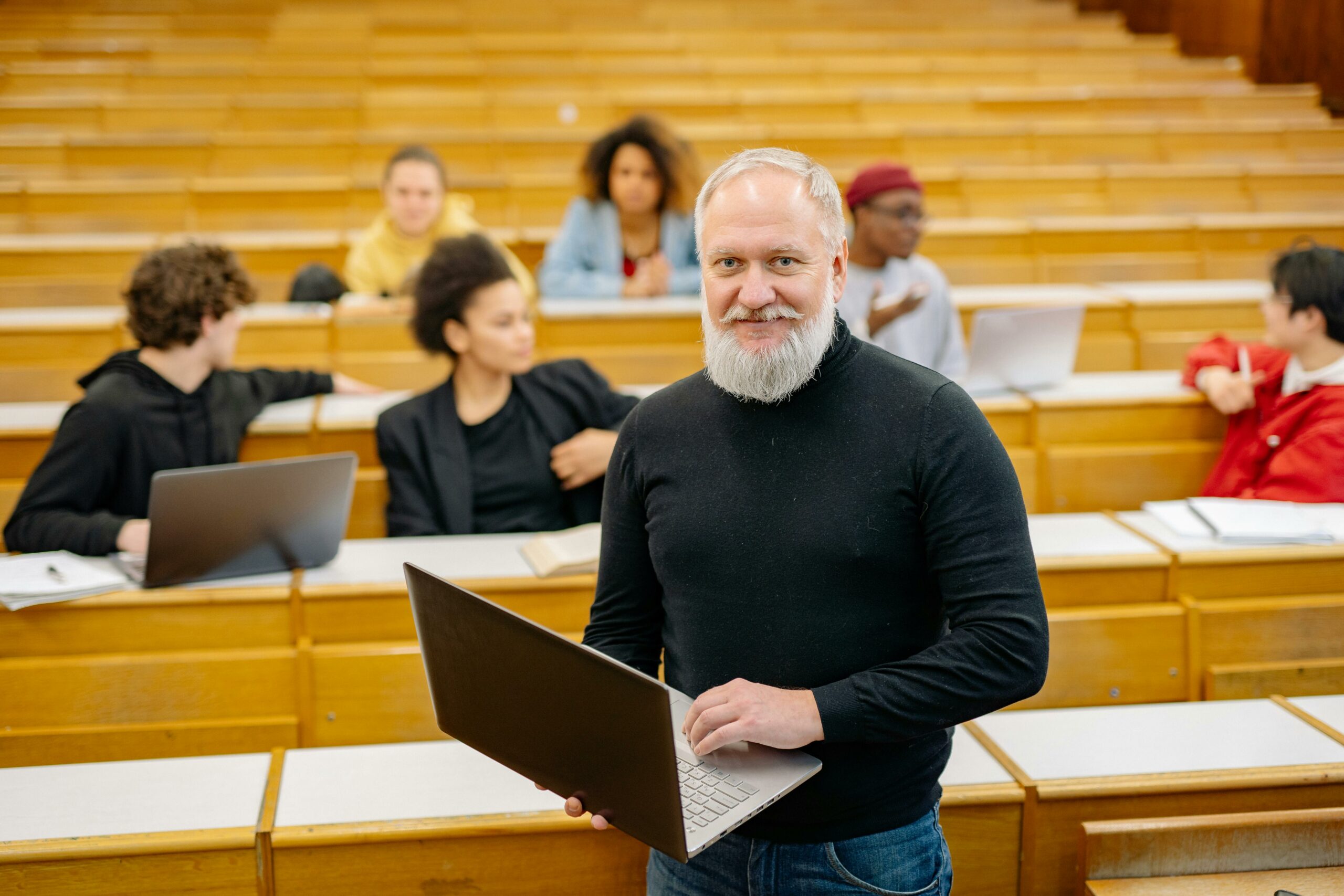 Professor standing in front of a lecture hall. He is holding a laptop and smiling at the camera.