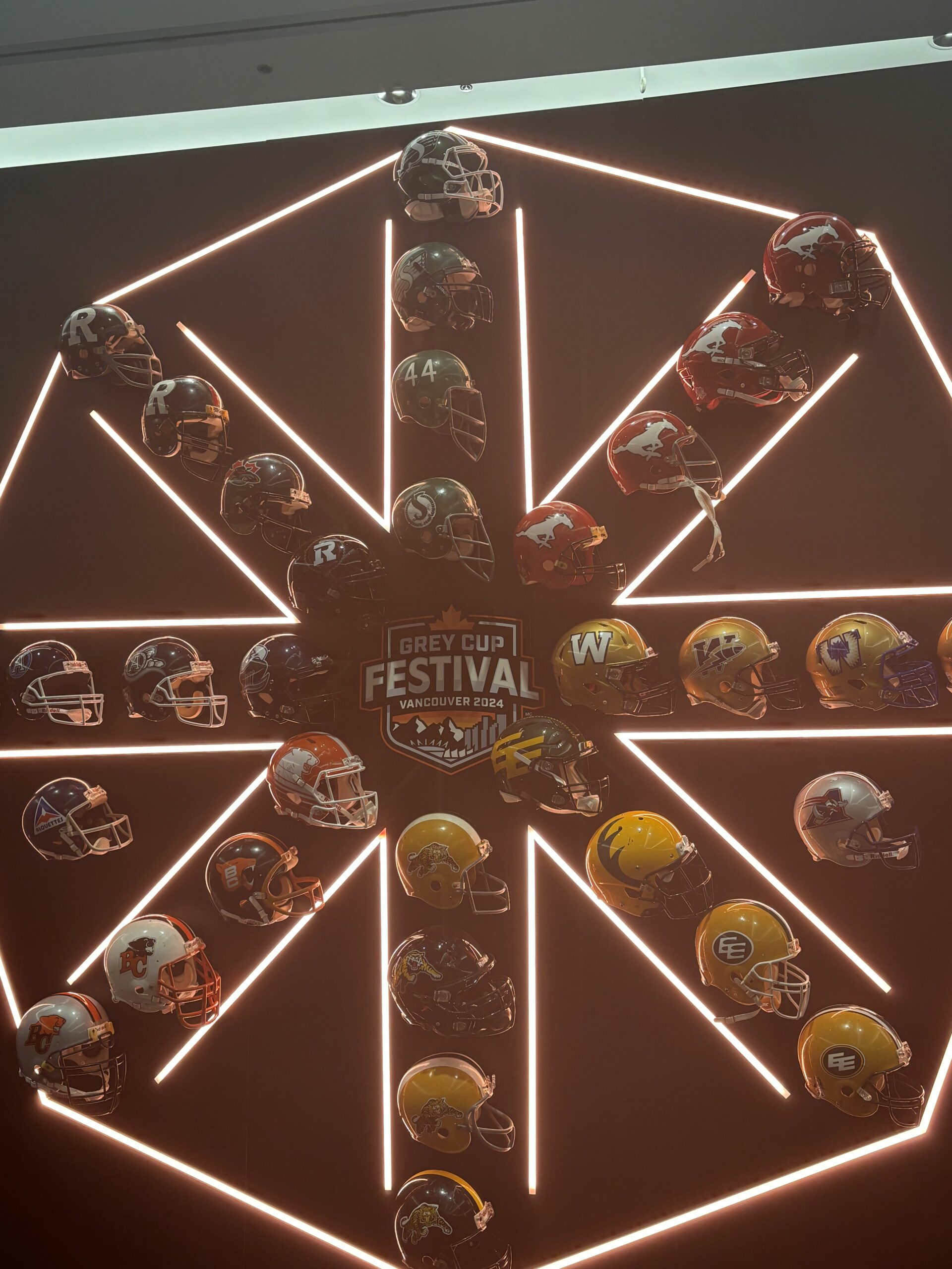 A wall of orange lights and helmets that form a snowflake-like shape. In the middle is a logo that reads “Grey Cup Festival Vancouver 2024.”