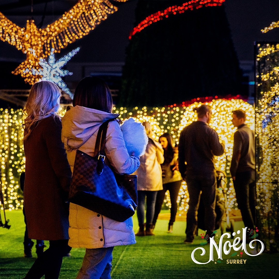 Two women with their backs to the camera wandering around an outdoor space filled with small, golden yellow lights.