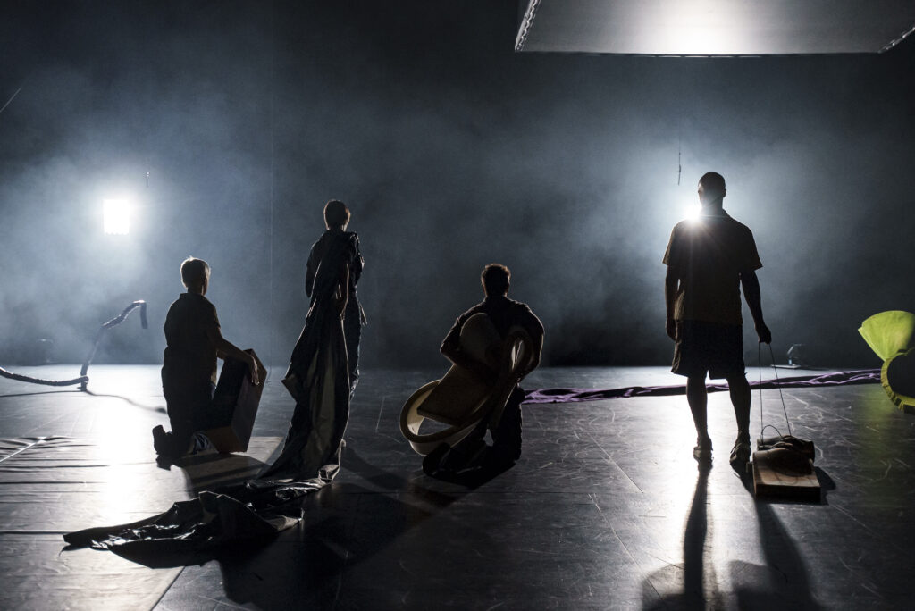 Four individuals pose with their backs to the camera, their figures illuminated by a light that shines from in front of them.