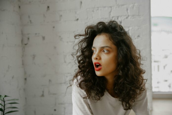 Astonished Woman with curly hair and red lipstick sitting in front of a white brick wall. She has her mouth open in shock.