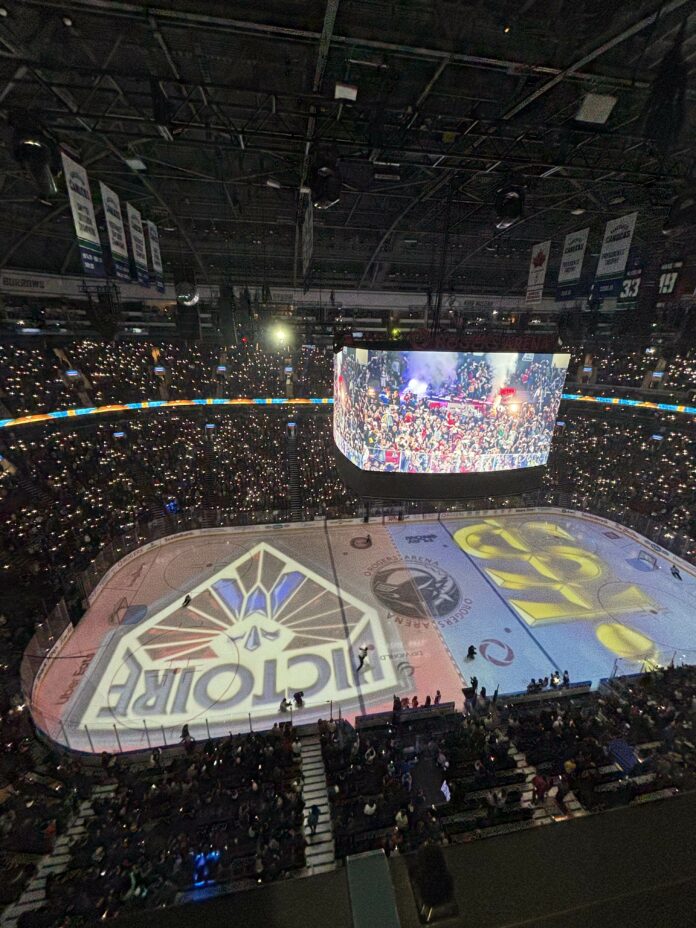 A hockey rink illuminated with individual lights. Displayed on the ice are a red, diamond-shaped logo with blue streaks, and a yellow T and S melded together against a blue background.