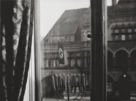 A black and white photograph of a lit menorah placed on a windowsill. Through the window, a building across the street with a flag of the Nazi regime.