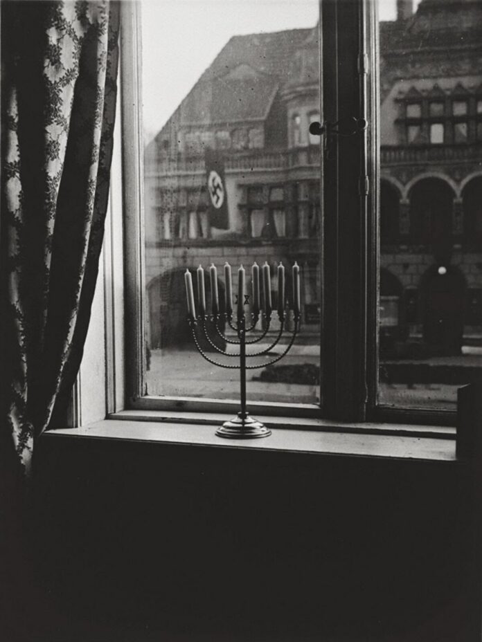 A black and white photograph of a lit menorah placed on a windowsill. Through the window, a building across the street with a flag of the Nazi regime.