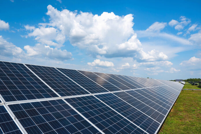 A row of solar panels in a field with clouds and a blue sky. Green energy production. This is a photo of a long row of utility-scale solar panels outside on a bright, sunny day.