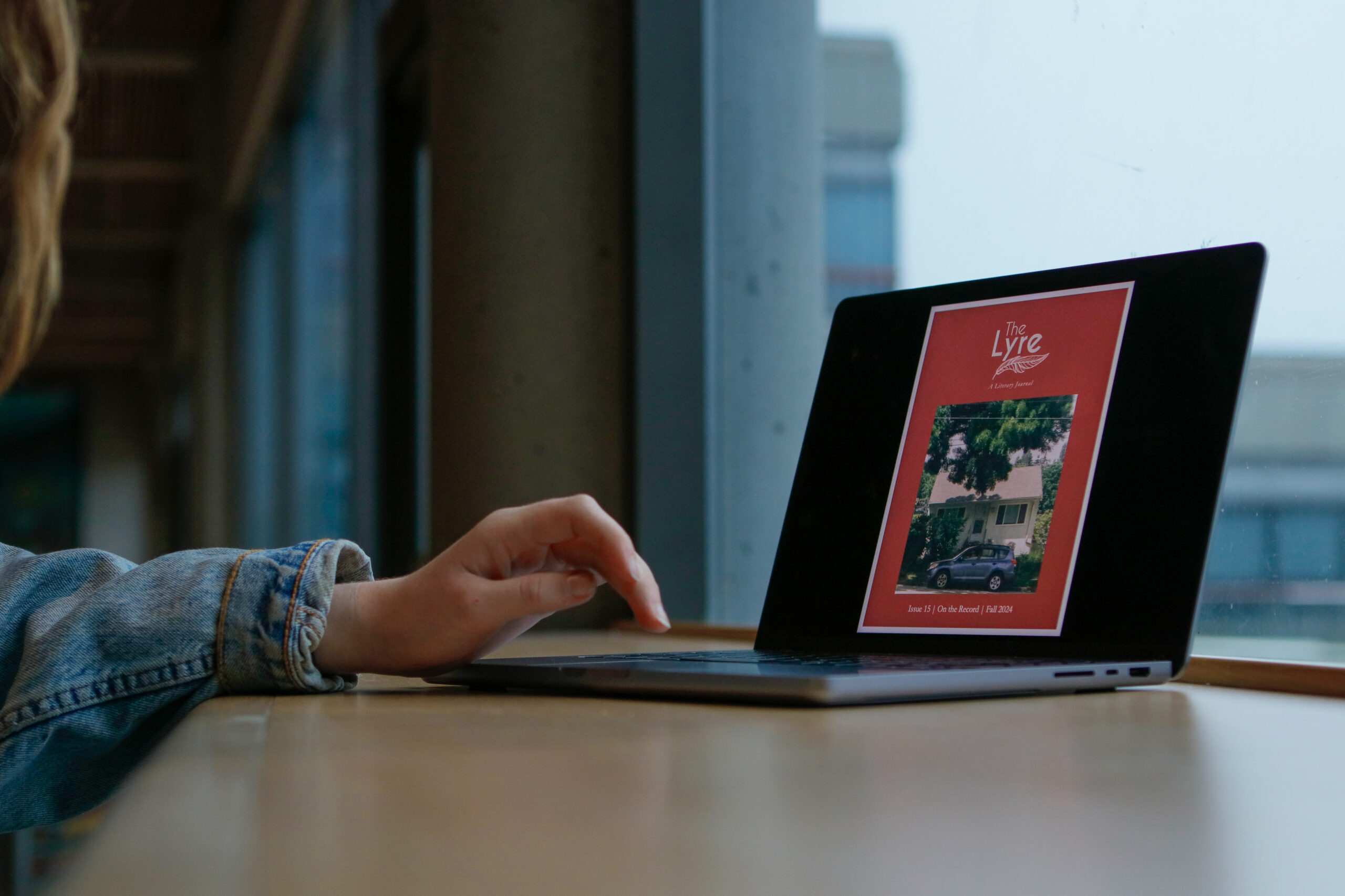 The cover of The Lyre’s 15th volume, which features the exterior of a house surrounded by shrubbery, is visible on a laptop screen situated near a window.