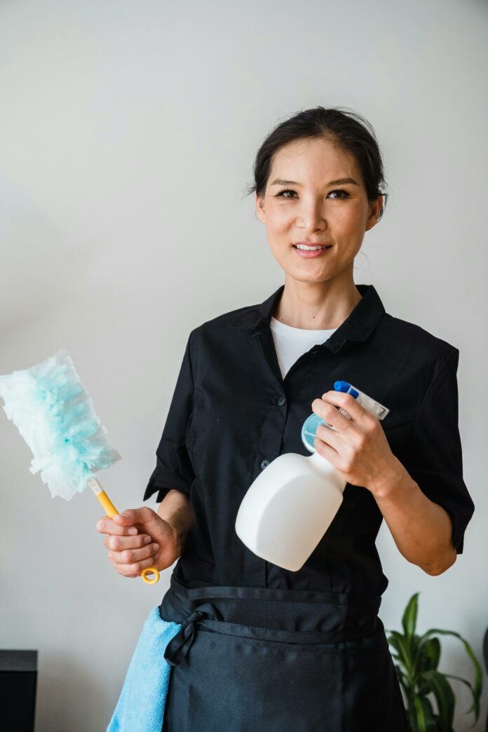 pexels-liliana-drew-9462192 Woman holding a spray bottle of cleaning solution and a duster. She has a cleaning rag in her pocket and is wearing a blue janitor jumpsuit.