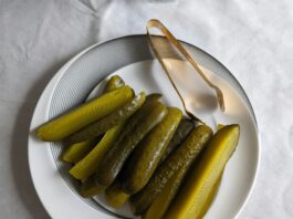 Pickle slices sit on a white plate beside a set of metal tongs.