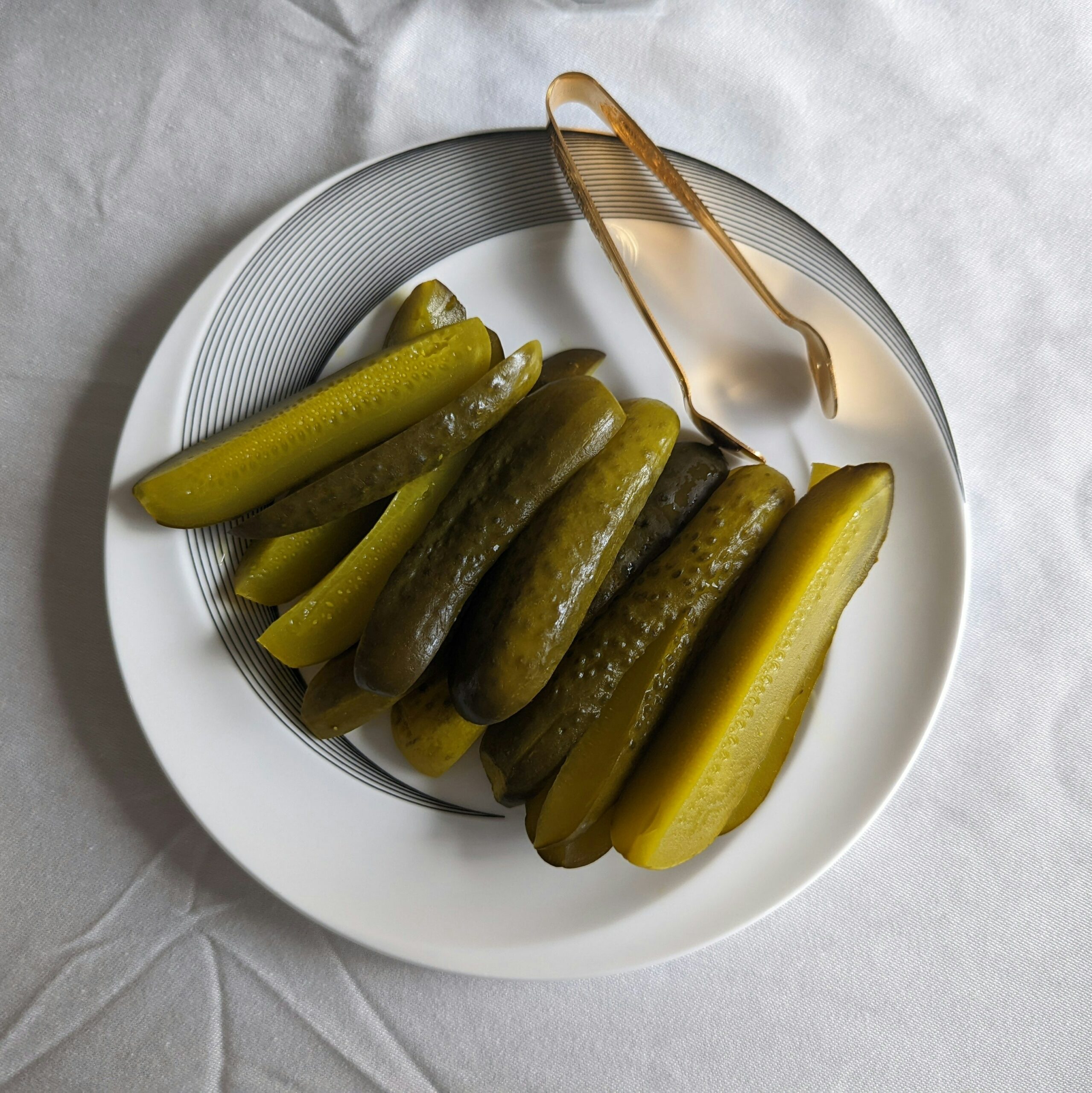 Pickle slices sit on a white plate beside a set of metal tongs.