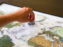 A hand reaches for a case of thumbtacks resting on a map of the world.