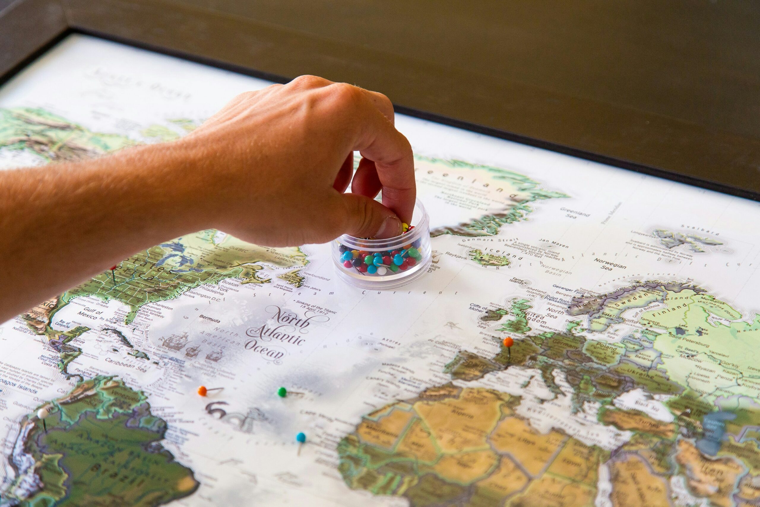 A hand reaches for a case of thumbtacks resting on a map of the world.