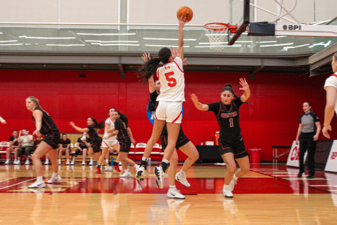 A girl in a white and red uniform attempts a lay-up while surrounded by players in black and red uniforms.