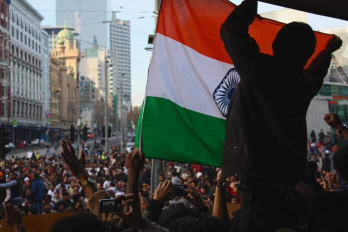 this picture is of a large group of students protesting in the street. A young man in the foreground holds the Indian flag up in front of him.
