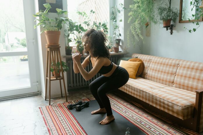 Mobility Woman in black workout clothes, squatting in her living room. She is surrounded by plants and she looks very focused.