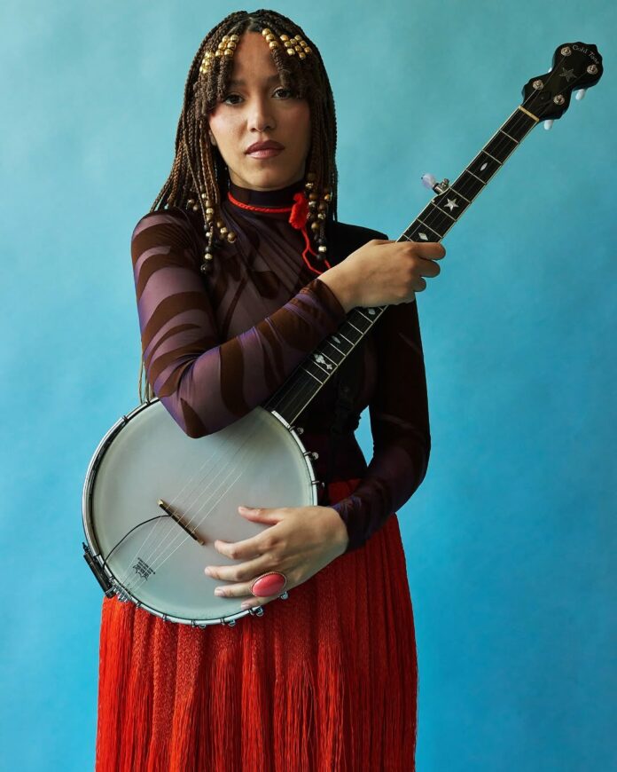 A girl with beaded dreadlocks holds a banjo in front of a blue background.