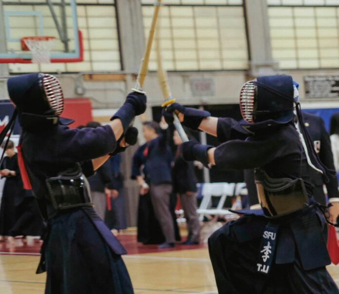 Two members of SFU’s kendo team, dressed in uniforms and wielding shinai, engage in combat with one-another.