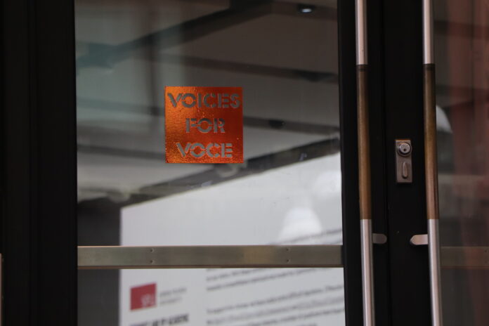 This is a photo of a shimmery orange window decal that reads “VOICES FOR VOCE” up on a door to a room that has an SFU presentation up.