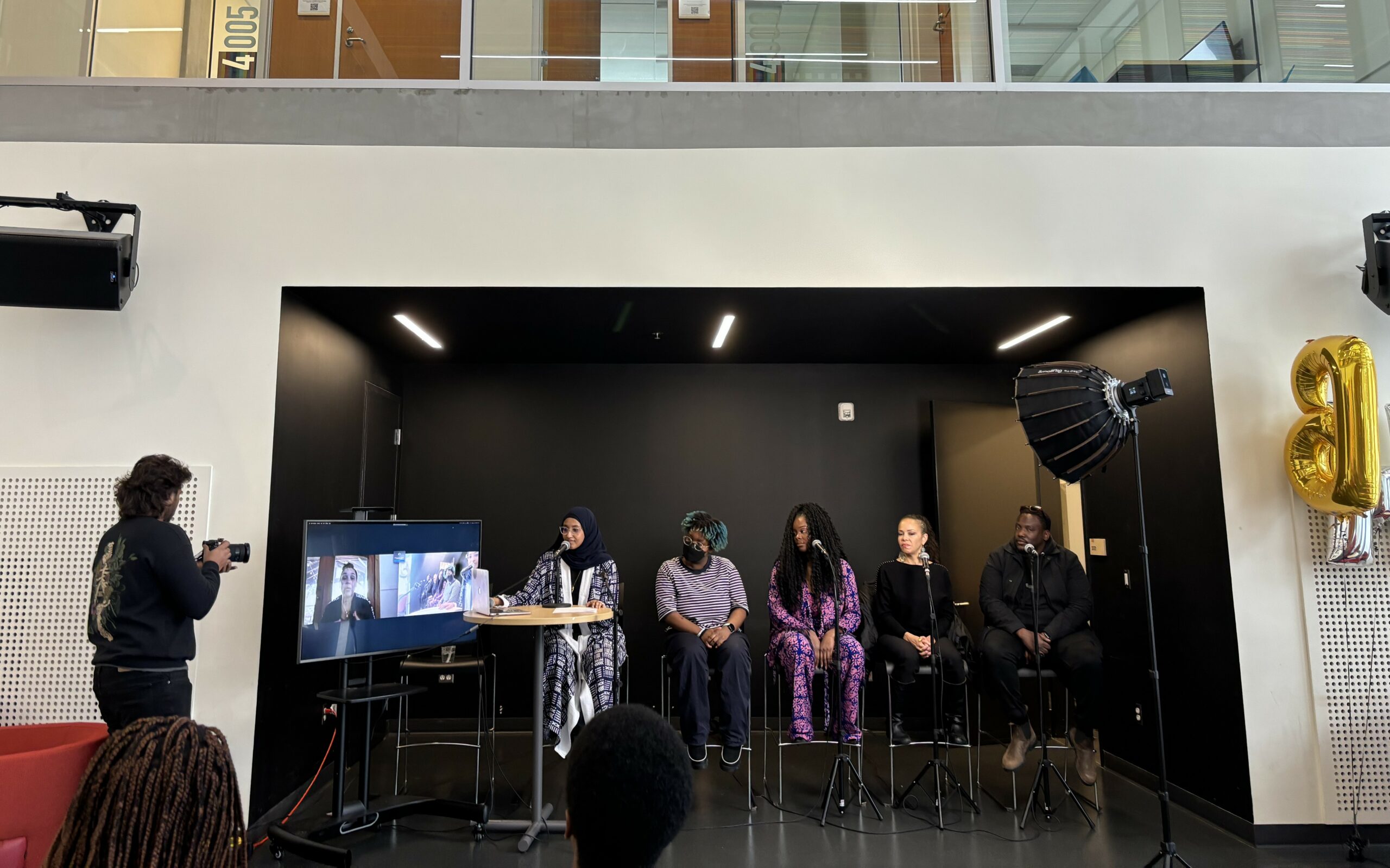 This is a photo of five black panelists sitting on high chairs. Some of them are looking over at a screen, where another panelist has joined them virtually.