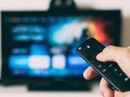 A person holds up a TV remote. In the background is a TV screen with a streaming service on it.