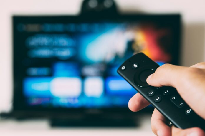 glenn-carstens-peters-EOQhsfFBhRk-unsplash A person holds up a TV remote. In the background is a TV screen with a streaming service on it.