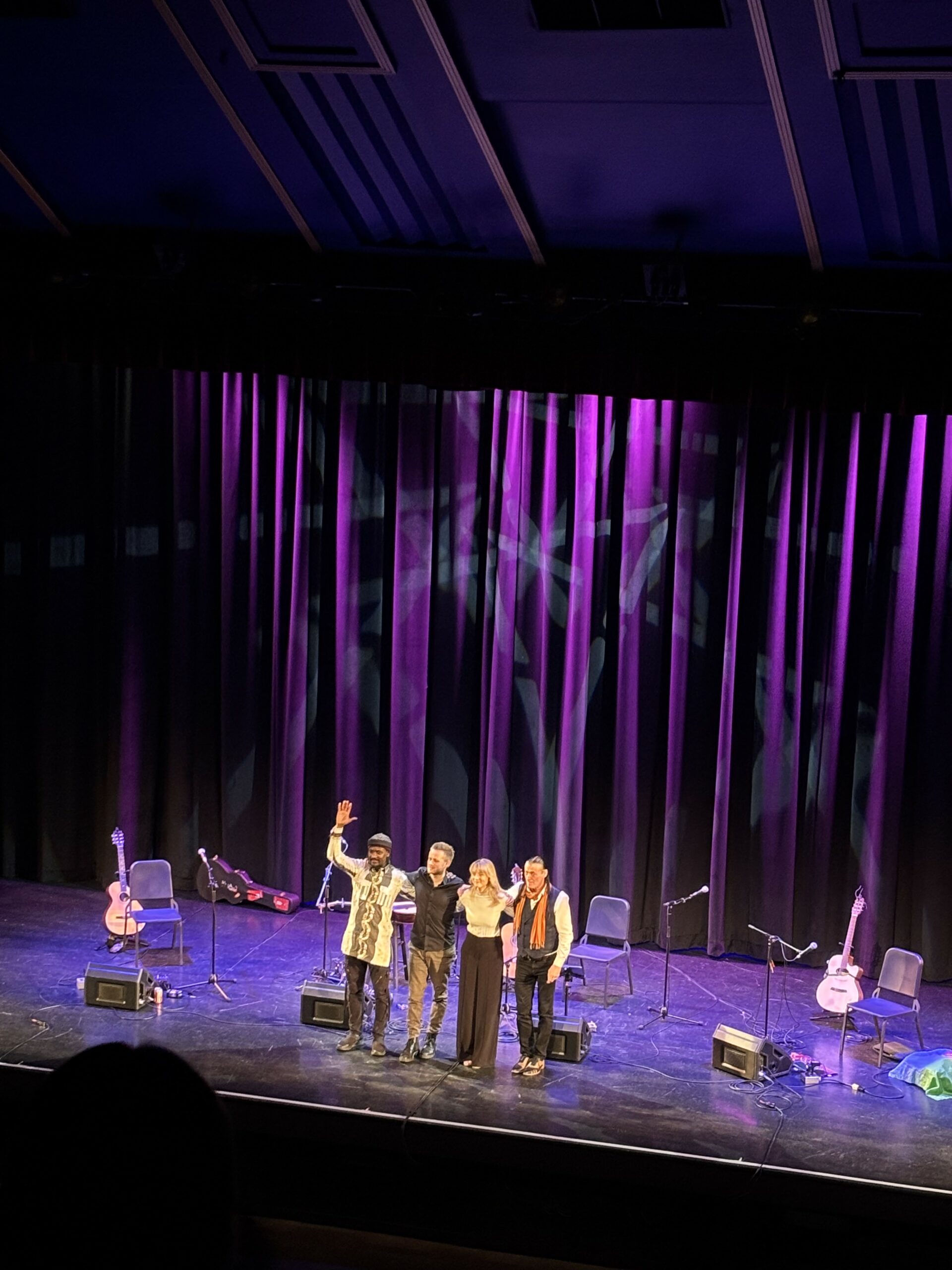 Four performers wave out to a stage while lit up in purple spotlights.