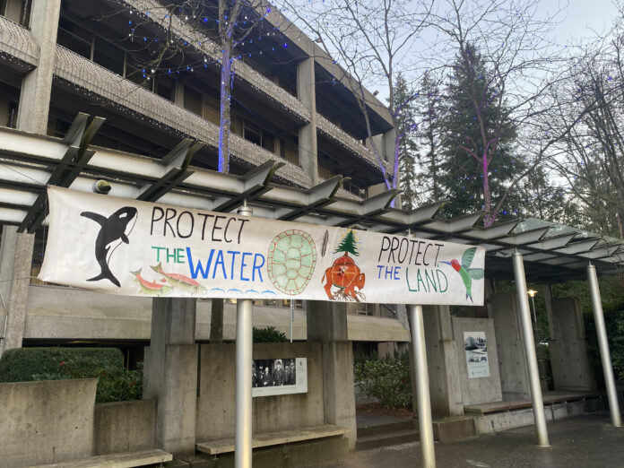 This is a photo of a hung-up banner that says “Protect the Water” / “Protect the Land.” It features drawings of an orca, salmon, a turtle shell, a bison, a hummingbird, and more.
