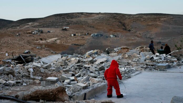 A small figure in all red stands amid concrete rubble and barren, brown hills.