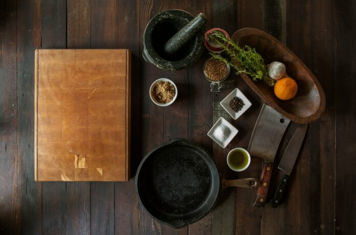 A wooden chopping board, black mortar and pestle, various spices, knives, and a cast-iron skillet on a wooden surface.