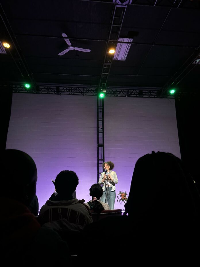 IMG_4722 A woman stands at a microphone while lit up by purple spotlights.