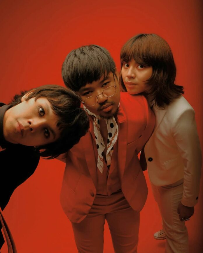 Three people stare at the camera wearing funky, 70s-esque suits against a deep red background.