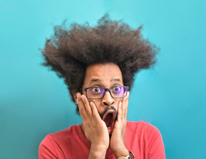 Shook Man looking shocked. He is wearing a red t-shirt, and black-framed glasses. He has his hands on his face and his mouth wide open. He has big eyes.