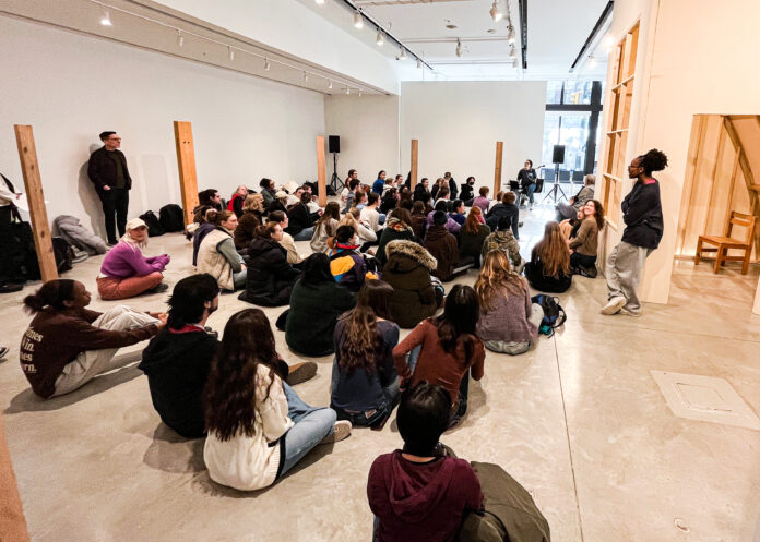 This is a photo of about 50 or 60 students sitting on the ground not facing the camera as they look toward someone distant at the front of the room. The room is very white, with a few wooden planks standing up, suggesting it is an arts showroom.