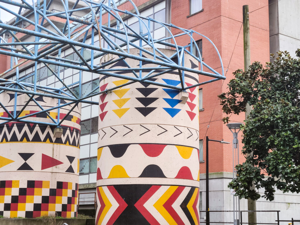 Two concrete pillars are decorated with red, black, blue, and yellow triangles arranged in downward-pointing columns. Red, black, and yellow swirly and triangular patterns wrap around the pillars in thick bands.