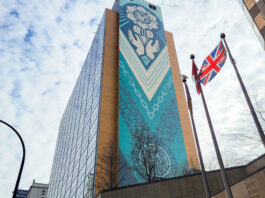 A blue, turquoise, and off-white mural lines one side of a tall building. Painted on the wall is a globe held up by two hands, surrounded by flower-like petals.