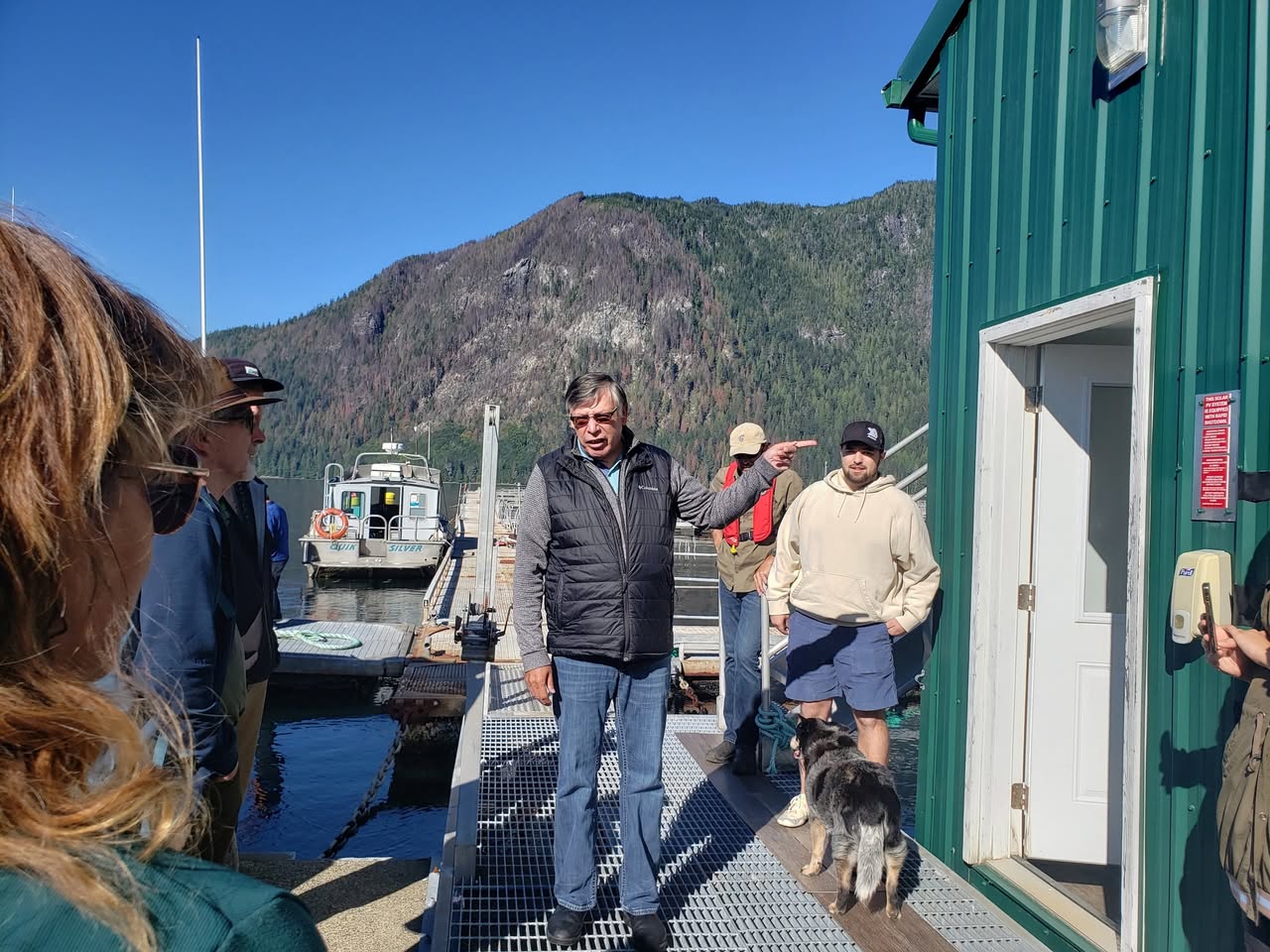 This is a photo of a man standing on a dock, pointing somewhere with his left hand as he talks to several other people. A mountain in the background and a multicoloured dog are also pictured.